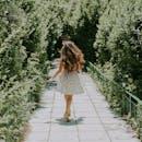 Back view of a woman in a sunlit park walking amidst lush green trees on a paved pathway.
